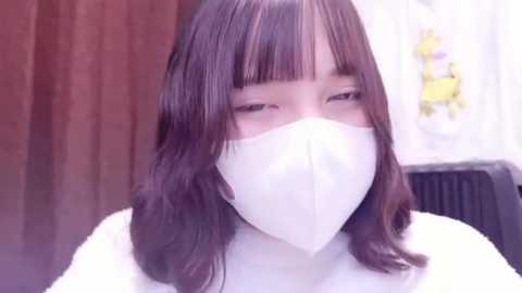 Media: Video of an Asian woman with shoulder-length brown hair and bangs, wearing a white mask and white top. Background shows a blurred, beige curtain and a white floral-patterned fabric.