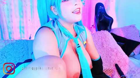 Media: Video of a woman with short, teal hair, wearing a sleeveless top, revealing large breasts. She's smiling, in a pink-lit room, leaning against a plush chair, with a watermark.