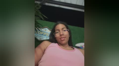 Video of a Latina woman with dark skin and long black hair, wearing glasses and a pink tank top, posing indoors with a green wall and a vintage sign in the background.