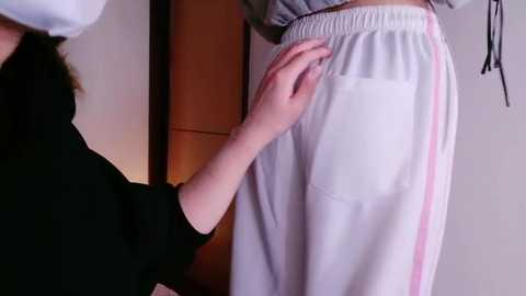 Media: Video of a person wearing a white sweatshirt with pink stripes and white sweatpants. A hand is adjusting the sweatpants pocket. Background shows a wooden door and a light-colored wall.