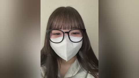 Media: Video of an East Asian woman with long, dark hair, wearing black-rimmed glasses, a white surgical mask, and a light green shirt, standing against a beige wall.