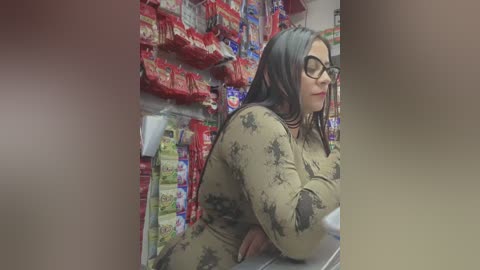 Video of a woman with long dark hair, wearing glasses, and a beige floral jumpsuit, bending over to check the bottom shelf of a grocery store aisle filled with colorful snack bags.