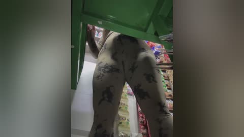 Video of a person with light skin wearing black and white patterned pants, bending over to lift a green object. Background shows shelves filled with colorful products.