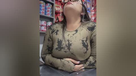 Media: Video of a woman with medium-length brown hair, wearing a tie-dye long-sleeve shirt, leaning back in a supermarket aisle filled with red and blue product displays.