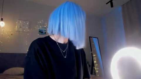 Media: A video of a person with a blue bob haircut, wearing a black sweater and necklace, standing in a dimly lit bedroom with a large circular mirror, fairy lights, and a bed visible in the background.