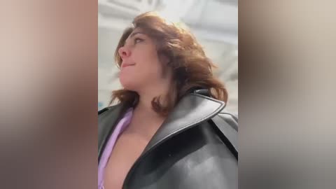 Media: Video of a woman with shoulder-length, wavy brown hair, wearing a black leather jacket over a purple shirt, looking upward with a slight smile. Background is blurred and white.