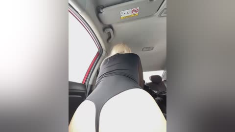 Media: A video from inside a car shows a blonde woman in a black leotard, leaning forward. Her exposed buttocks are prominent, highlighting her curves. The car's interior is visible, including a sign and a person sitting in the backseat.