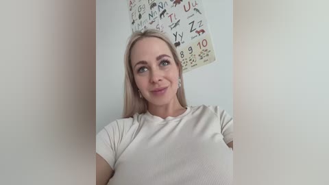 Media: A video of a smiling Caucasian woman with straight blonde hair, wearing a white ribbed shirt, standing against a white wall adorned with a colorful alphabet poster.