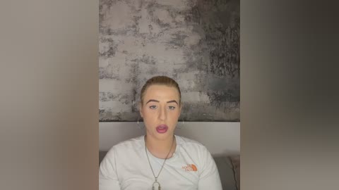 A video of a fair-skinned, blonde woman with a neutral expression, wearing a white long-sleeve shirt and a necklace, against a textured gray wall.