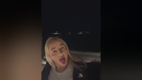 Media: Video of a blonde woman with glasses, wearing a black jacket, taken at night with a bright light source, creating a dramatic, slightly blurred background.