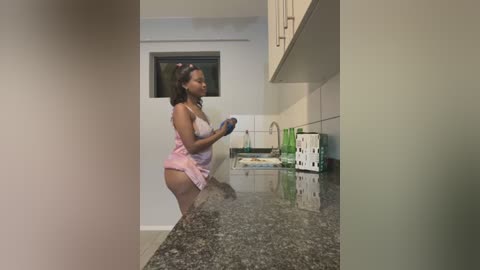 Media: Video of a pregnant Black woman in a pink apron, washing dishes in a modern kitchen with white walls and a black granite countertop.