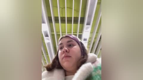 Media: Video of a young Caucasian woman with fair skin, long brown hair, and a neutral expression, lying in a white crib with vertical bars and a lime green cushion. The image is slightly blurred, giving a soft focus.