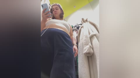 Media: A candid video of a plus-sized woman with medium skin tone, wearing a white crop top and blue sweatpants, taking a selfie in a brightly lit, modern bathroom with yellow ceiling panels.