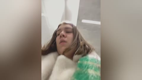 Media: A close-up video of a young woman with long, brown hair, eyes closed, and mouth slightly open, lying on a white bed, wearing a green and white checkered blanket.