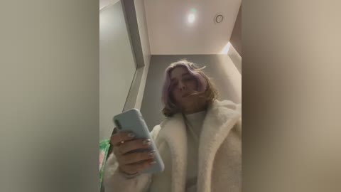 Media: Video of a woman with light skin and shoulder-length, pastel purple hair, wearing a white bathrobe, taking a selfie in a dimly lit bathroom.