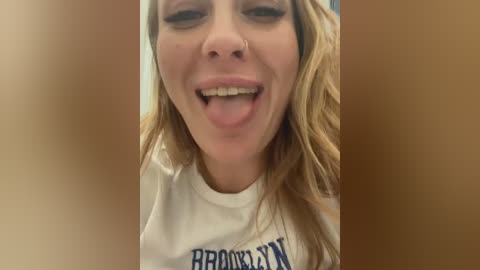 Media: Video of a smiling woman with light skin and long, wavy blonde hair, wearing a white \"Brooklyn Nets\" t-shirt. She sticks out her tongue playfully.