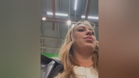 Media: Video of a young woman with long blonde hair, wearing a black leather jacket, standing in a warehouse with exposed pipes and green walls.