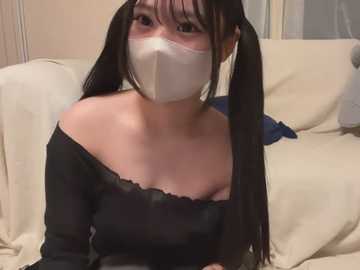 Media: Video of an Asian woman with long black pigtails, wearing a black off-shoulder top, white face mask, and seated on a beige couch. Background shows a beige wall and a white door.