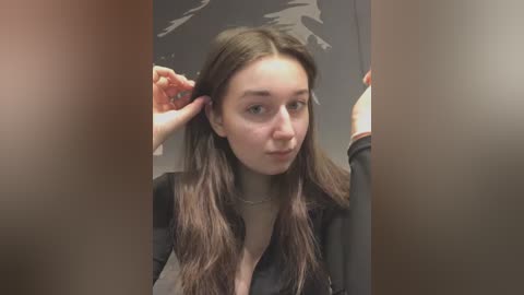 Media: Video of a young Caucasian woman with long brown hair, wearing a black shirt, adjusting her hair in a mirror. Background is a blurred, dark room with a faint wall art.