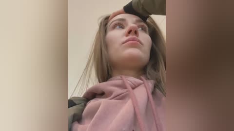 Media: Video of a young Caucasian woman with light skin, long brown hair, and blue eyes. She wears a pink hooded sweatshirt. A hand gently touches her forehead, indicating a moment of tenderness or reassurance.