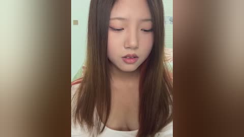 Media: Video of a young Asian woman with long, straight brown hair, wearing a white top, and light makeup, looking down with a neutral expression. Background is blurred, suggesting an indoor setting.