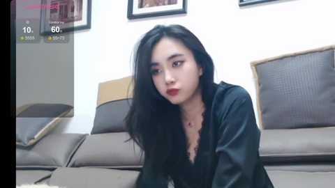 Media: A video of an East Asian woman with long black hair, fair skin, and red lipstick, wearing a black jacket, sitting on a beige sofa in a modern living room with framed pictures on the white wall.