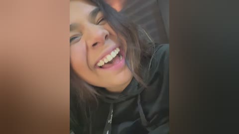 A close-up video of a young woman with medium brown skin, smiling widely, with long dark hair and a black jacket.