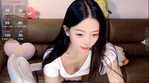 Media: Video of an Asian woman with long black hair, wearing a white top, lying on a brown leather couch. Background includes stuffed animals and a smartphone displaying a live stream.