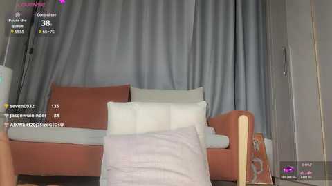 Media: Video of a cozy living room with a light gray curtain backdrop, a tan sofa, white pillows, and a light pink blanket.