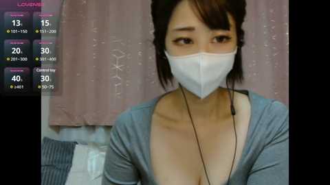 Media: Video of an East Asian woman with short black hair, wearing a grey cardigan and white mask, looking worried. Background features a pink curtain and a grey sofa.