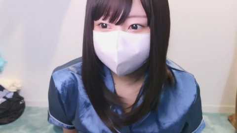 Video of an Asian woman with straight black hair, wearing a white mask and blue satin shirt, standing indoors with a blurred background.