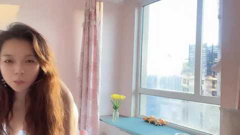 Media: A video of a young Asian woman with long brown hair, wearing a white top, standing near a window in a brightly lit room with a view of an urban cityscape.