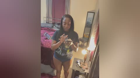 A video of a young woman with dark skin and shoulder-length hair, wearing a dark t-shirt and shorts, standing in a modest bedroom with a bed, mirror, and lamp.