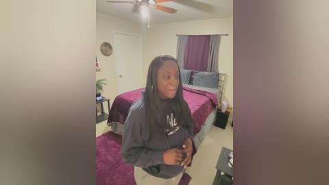 Video of a young Black woman with braids in a dark blue sweater standing in a dimly lit bedroom with a maroon bedspread, white walls, and a ceiling fan.