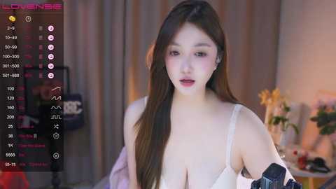 Media: Video of a young Asian woman with long brown hair, fair skin, wearing a white bra, and a black strap-on. Background shows a dimly lit bedroom with a bed and a plant.