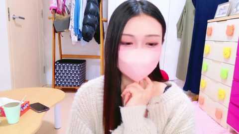 Media: Video of an Asian woman with long black hair, wearing a white mask, sitting in a brightly lit room with a wooden table, hanging clothes, and a colorful dresser.