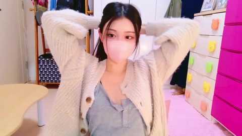 Media: Video of an East Asian woman with long black hair, wearing a white mask, beige cardigan, and light blue top, sitting with hands behind head in a colorful, cluttered room.