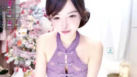 Media: A video of an East Asian woman with fair skin and dark hair in a purple, lace halter top, smiling softly in a cozy, festive room with a Christmas tree in the background.