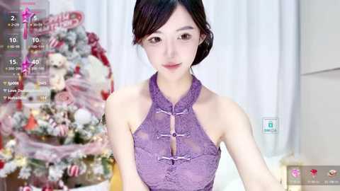 Media: A video of an East Asian woman with fair skin, wearing a purple, lace-trimmed halter top, standing in front of a decorated Christmas tree.