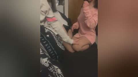 Media: Video of a young Asian woman with fair skin, sitting on a bed, wearing a pink sweater and black leggings. A man in a white sweater is touching her face.