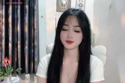 Media: A video of an East Asian woman with long black hair, fair skin, and red lipstick, wearing a white top, sitting indoors. The background includes a potted plant and curtains.