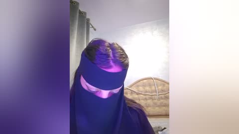 Media: Video of a person in a dark blue hijab, standing against a light purple wall with a white textured backdrop and a wicker headboard, creating a surreal, mysterious atmosphere.