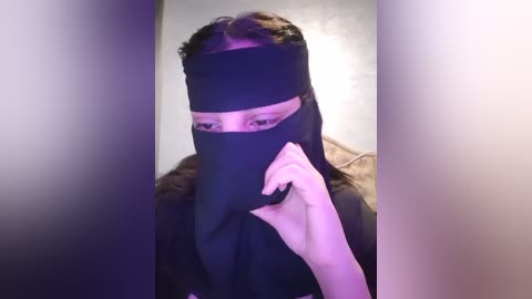 Media: A video of a person wearing a black niqab, which covers the face except for the eyes, holding a phone. The background is blurred, with a hint of a beige wall and a purple light.