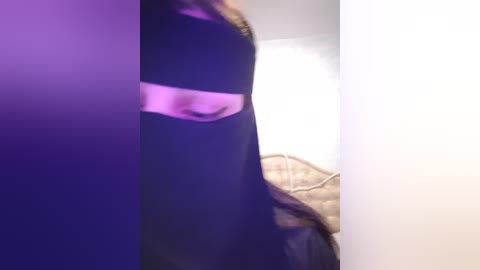 Media: A blurry, low-resolution video captures a woman in a black dress with a white sash, standing beside a bed with white sheets and a beige blanket.