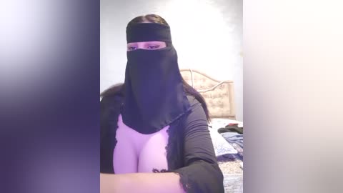 Media: Video of a woman in a black niqab with pink glasses, wearing a black jacket over a light pink top, standing in a cluttered bedroom with a bed and scattered clothes.