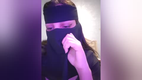 Media: A video of a person with light skin, wearing a black face mask and black top, standing against a blurred background. The person is holding their hand to their mouth, partially obscuring their face.