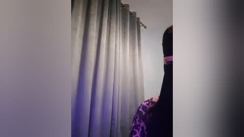 Media: A video captures a person with long, dark hair partially covered by a black mask, standing in a room with gray curtains and a leopard print blanket. The image is dimly lit with a soft, purple glow.