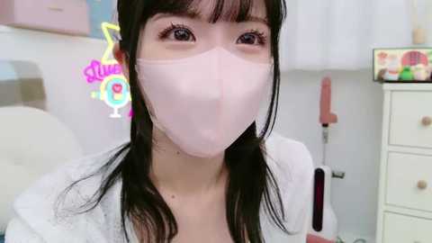 Media: Video of an Asian woman with long black hair, wearing a pink face mask and a white blouse, in a brightly lit room with neon signs and a white dresser in the background.