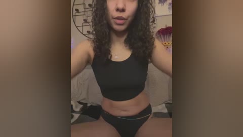 Media: A video of a young woman with curly dark hair, wearing a black sports bra and matching panties, sitting on a bed with a colorful wall and decorative items in the background.