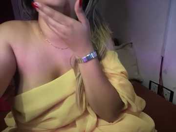 Media: A video of a fair-skinned woman with blonde hair, wearing a yellow off-shoulder dress and a silver bracelet, covering her mouth with her hand. The background is dimly lit with a hint of red and white furniture.